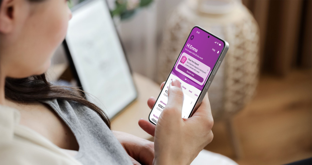 Woman holding a phone showing the AIB Mobile Hi Emma home screen
